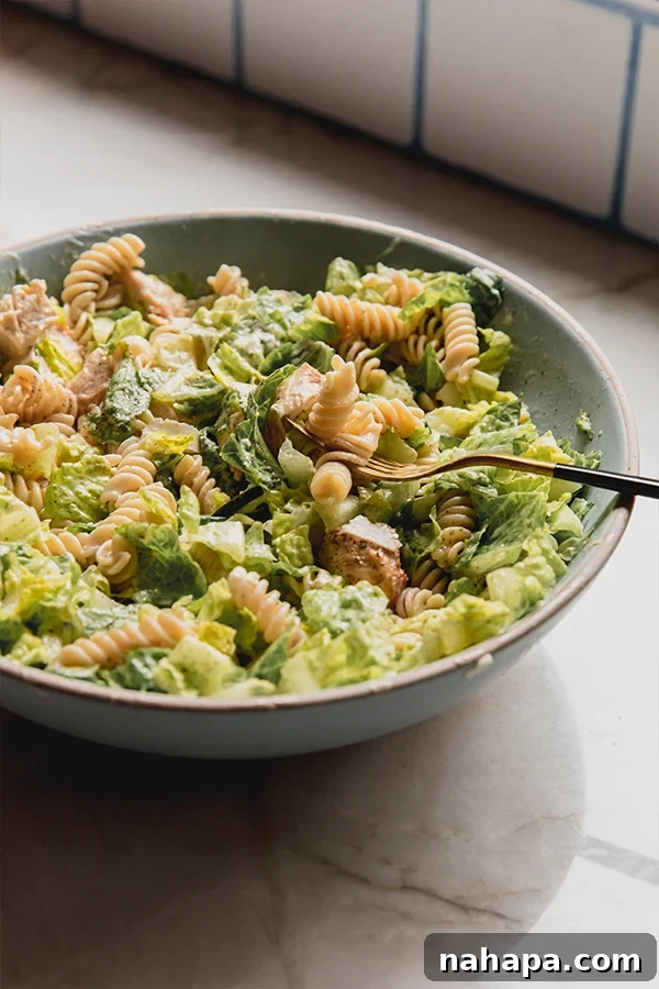 A close-up shot of a bowl of chicken caesar pasta salad with a fork inside, ready to eat.
