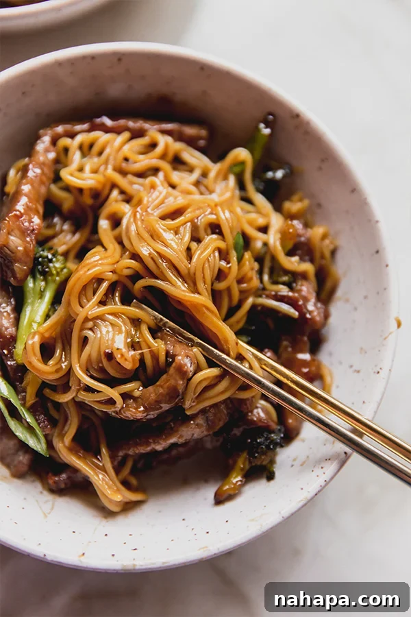 A bowl of sheet pan Mongolian beef in a bowl with chopsticks taking a bite out of it, showcasing the rich sauce and fresh green onion garnish.