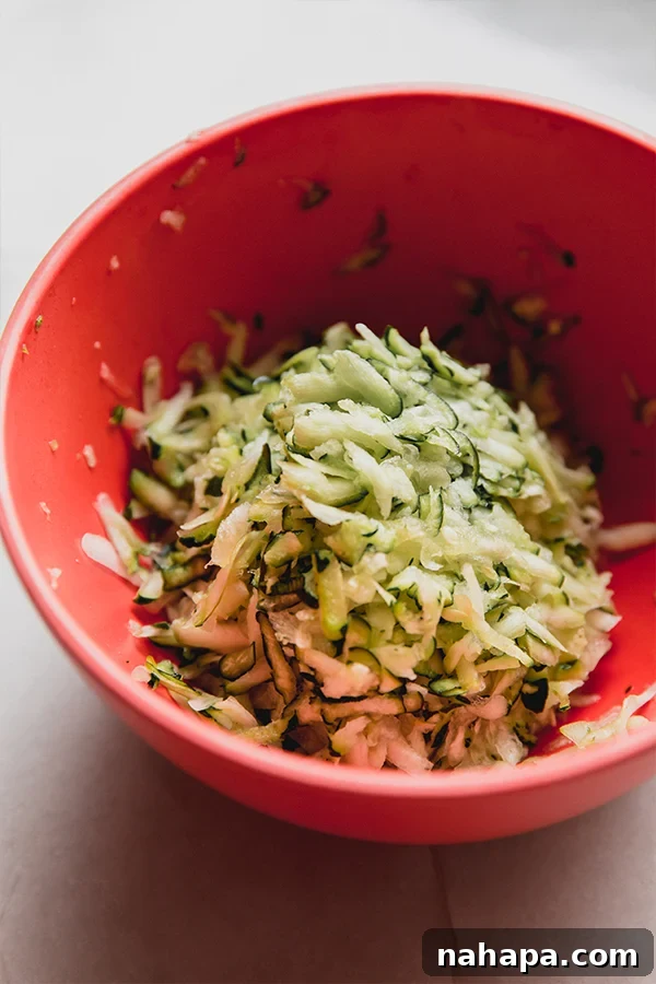 A close-up shot of a mixing bowl filled with freshly shredded zucchini, showing its fine texture and vibrant green color before being incorporated into the dish.