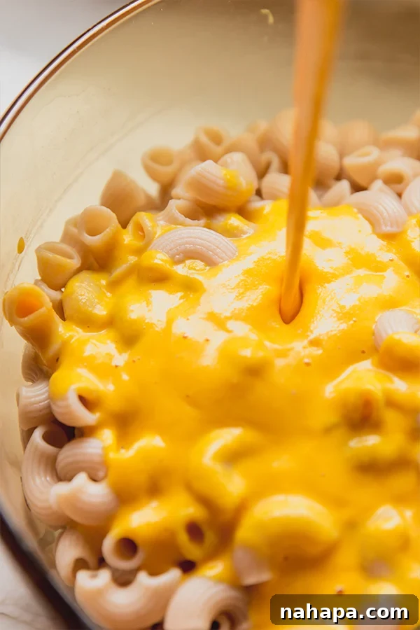 A pot of cooked elbow pasta on a stovetop, with a rich, creamy orange sauce being poured over it, preparing for the final combination of ingredients.