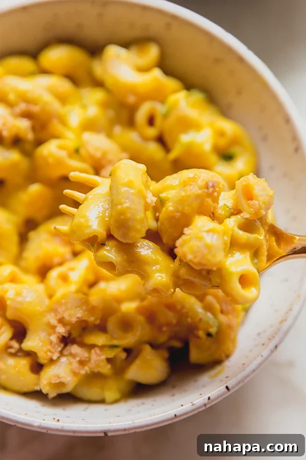 A close-up of a fork taking a bite from a bowl of zucchini mac and cheese, highlighting the creamy texture and the golden brown topping, inviting viewers to taste.