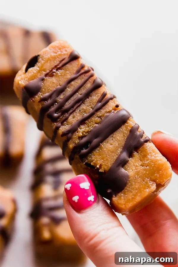 A hand holding a single homemade cookie dough protein bar, showcasing its texture and the chocolate drizzle on top, ready to be eaten.