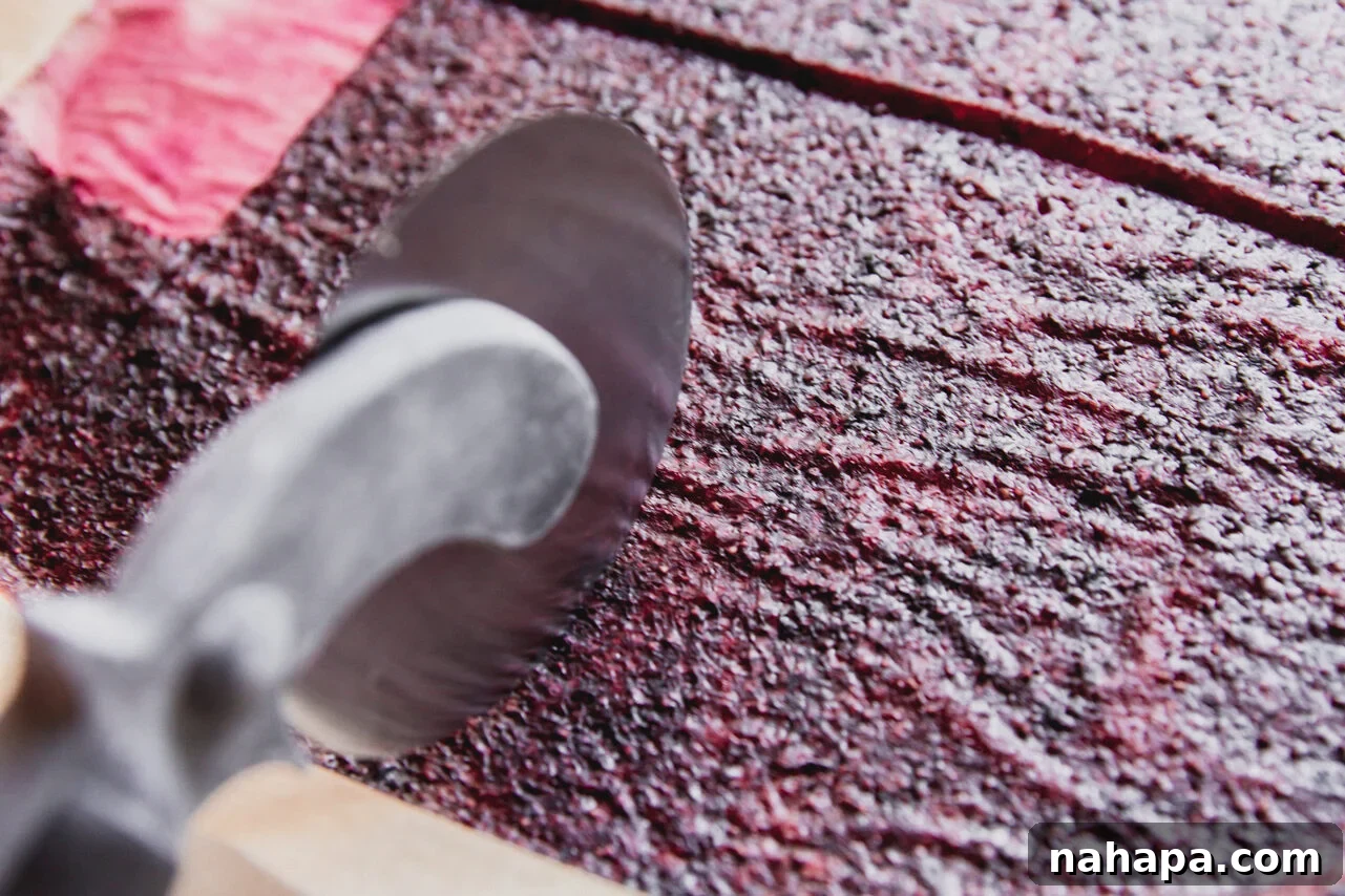A sheet of cooled, homemade fruit leather being precisely sliced into individual, uniform strips using a pizza cutter on a clean surface.