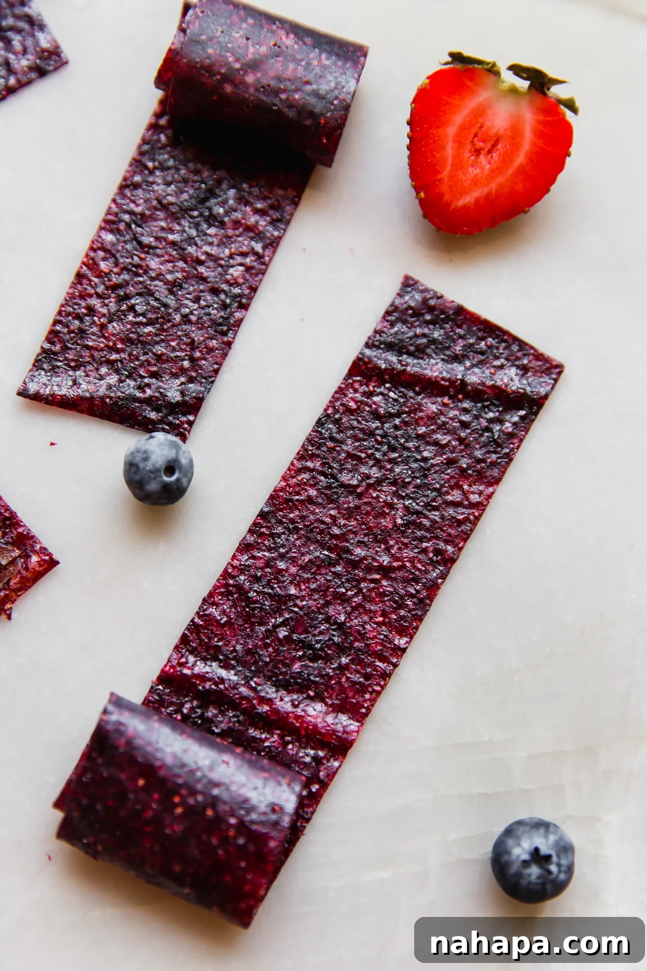 Assortment of vibrant homemade fruit roll-ups on a white background, beautifully arranged with fresh strawberries and blueberries.