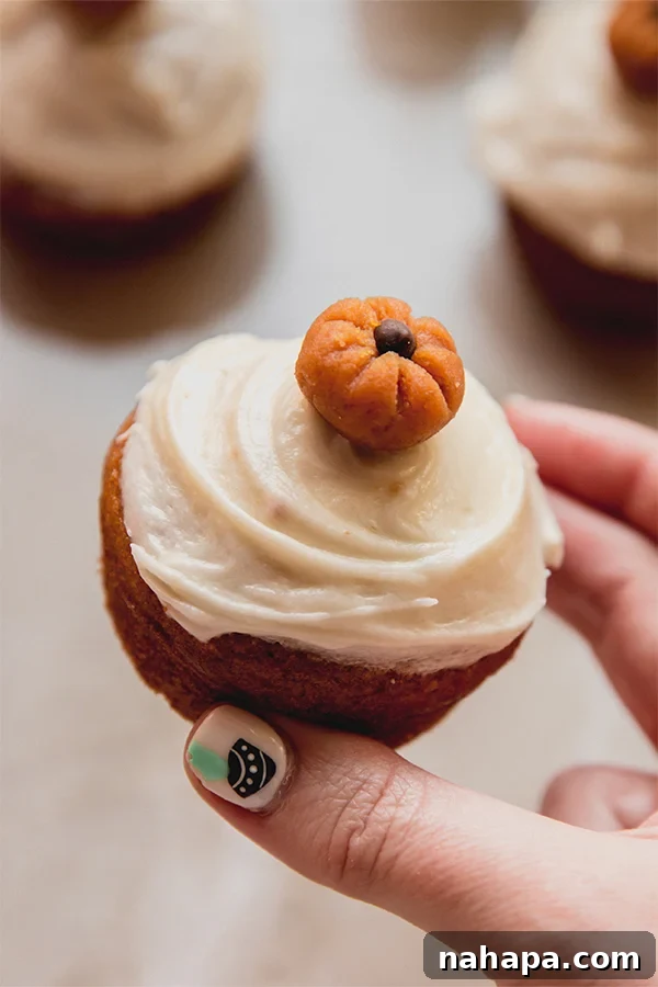A pumpkin spice cupcake being held, decorated with cream cheese frosting and a mini pumpkin buckeye.