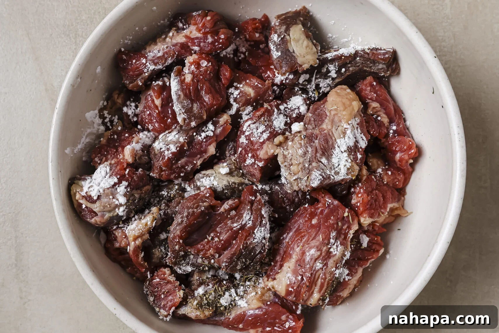 Beef stew meat being tossed with arrowroot starch and seasoning in a bowl.