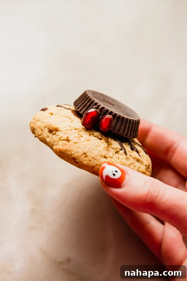 A close-up of a decorated Halloween spider cookie being held in a hand, showing the detailed almond butter cup body, chocolate legs, and candy eyes.