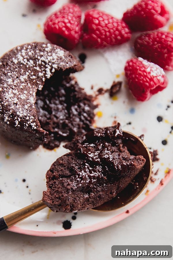 A chocolate lava cake on a plate with a spoon taking a bite out of the cake, showing the molten center, accompanied by fresh raspberries.