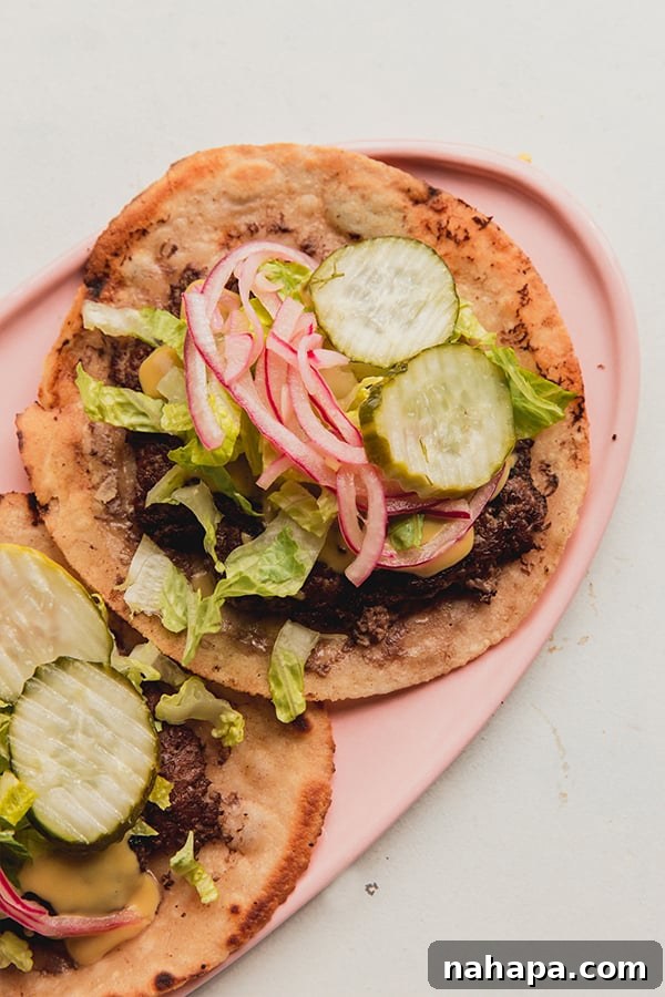 A serving plate with two smash burger tacos topped with pickled onions and pickles, looking incredibly appetizing.