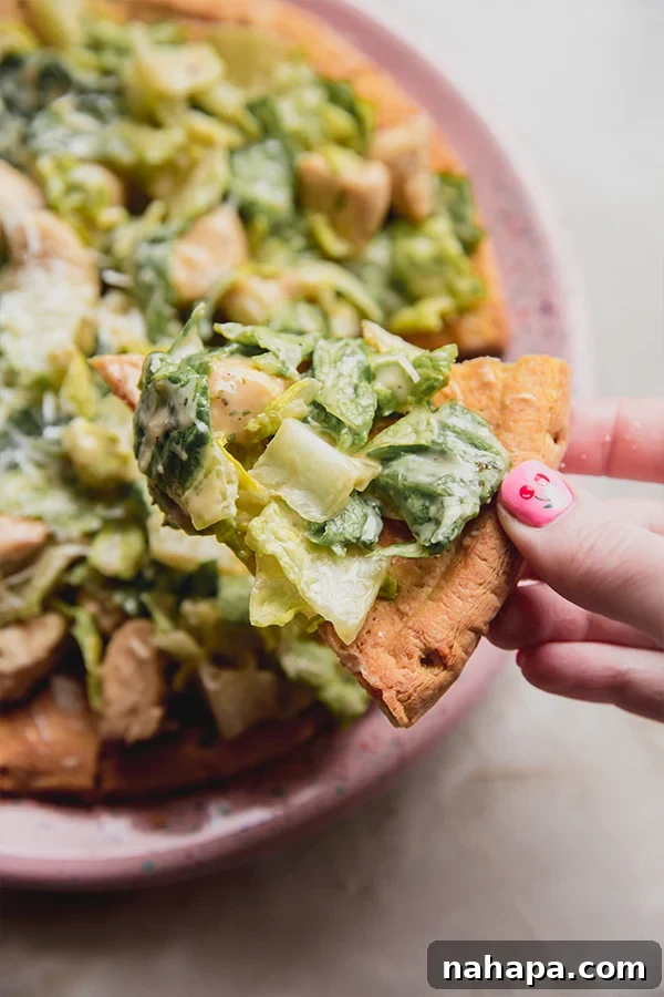 A piece of chicken caesar salad pizza being taken from the pizza, showing the crisp crust and fresh toppings.