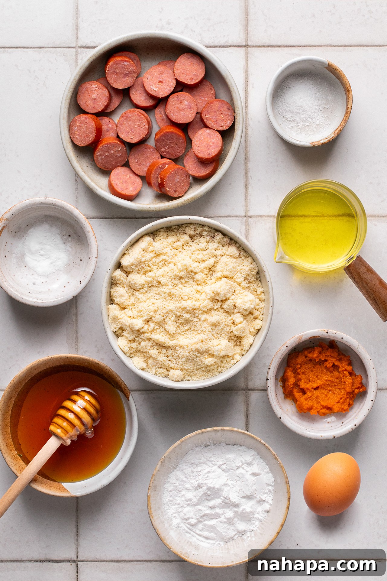 A collection of ingredients for corn dog muffins, including almond flour, tapioca starch, hot dogs, pumpkin puree, eggs, avocado oil, and honey.