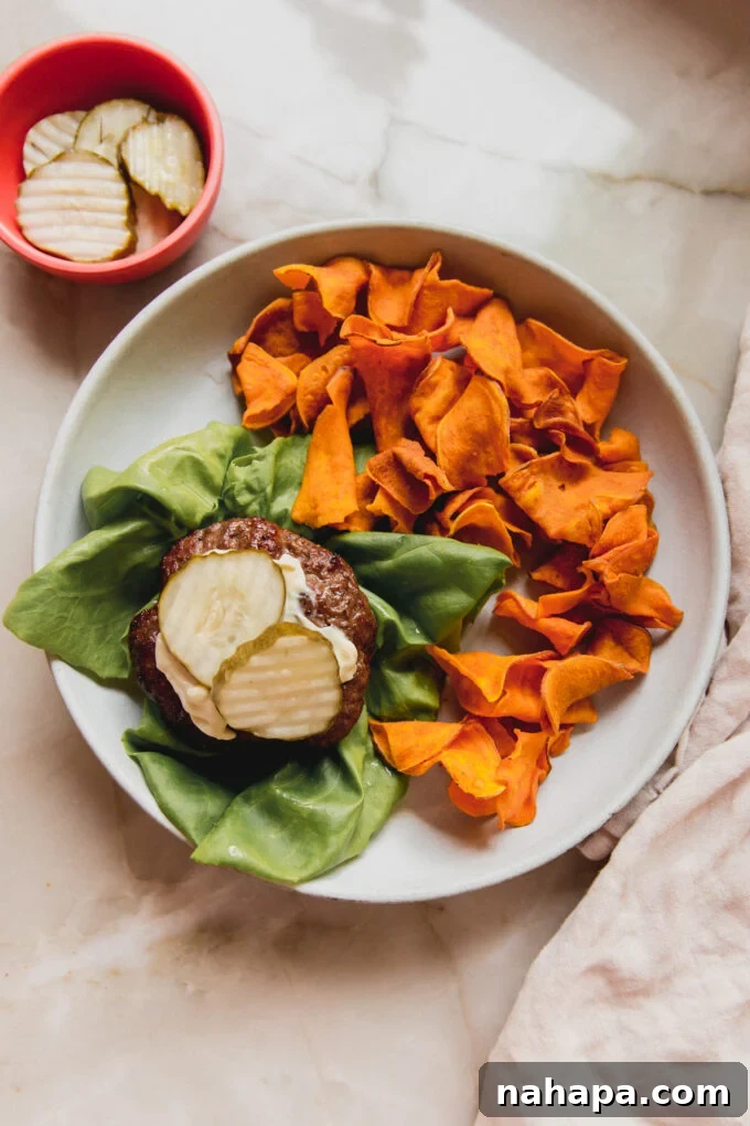 Air fryer burger artfully arranged on a crisp lettuce wrap, adorned with creamy mayonnaise and crunchy pickles, served alongside a side of golden sweet potato chips.