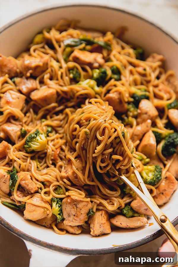 Tender chicken and crisp broccoli ramen noodles beautifully coated in a savory sauce, being tossed with tongs in a pan, ready to be served.