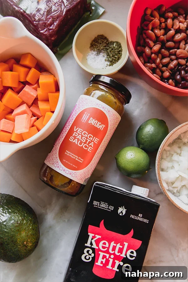 An overhead photo displaying the core ingredients for nightshade-free chili: ground beef, beans, limes, diced onion, a jar of nightshade-free pasta sauce, beef broth, and sliced avocado.