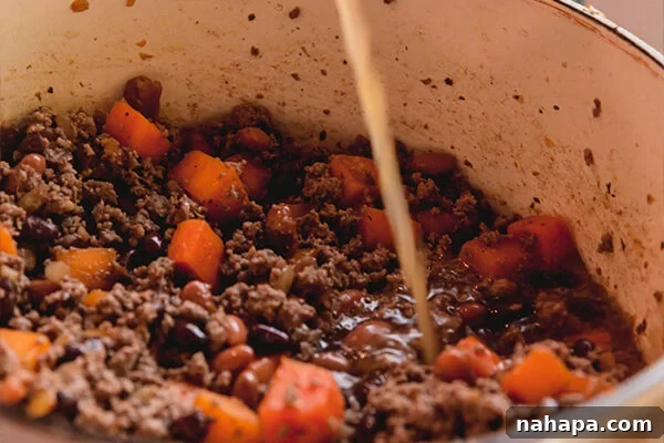 Sweet potato, onion, and beans simmering in a pot as beef broth is poured in, combining all the chili components.