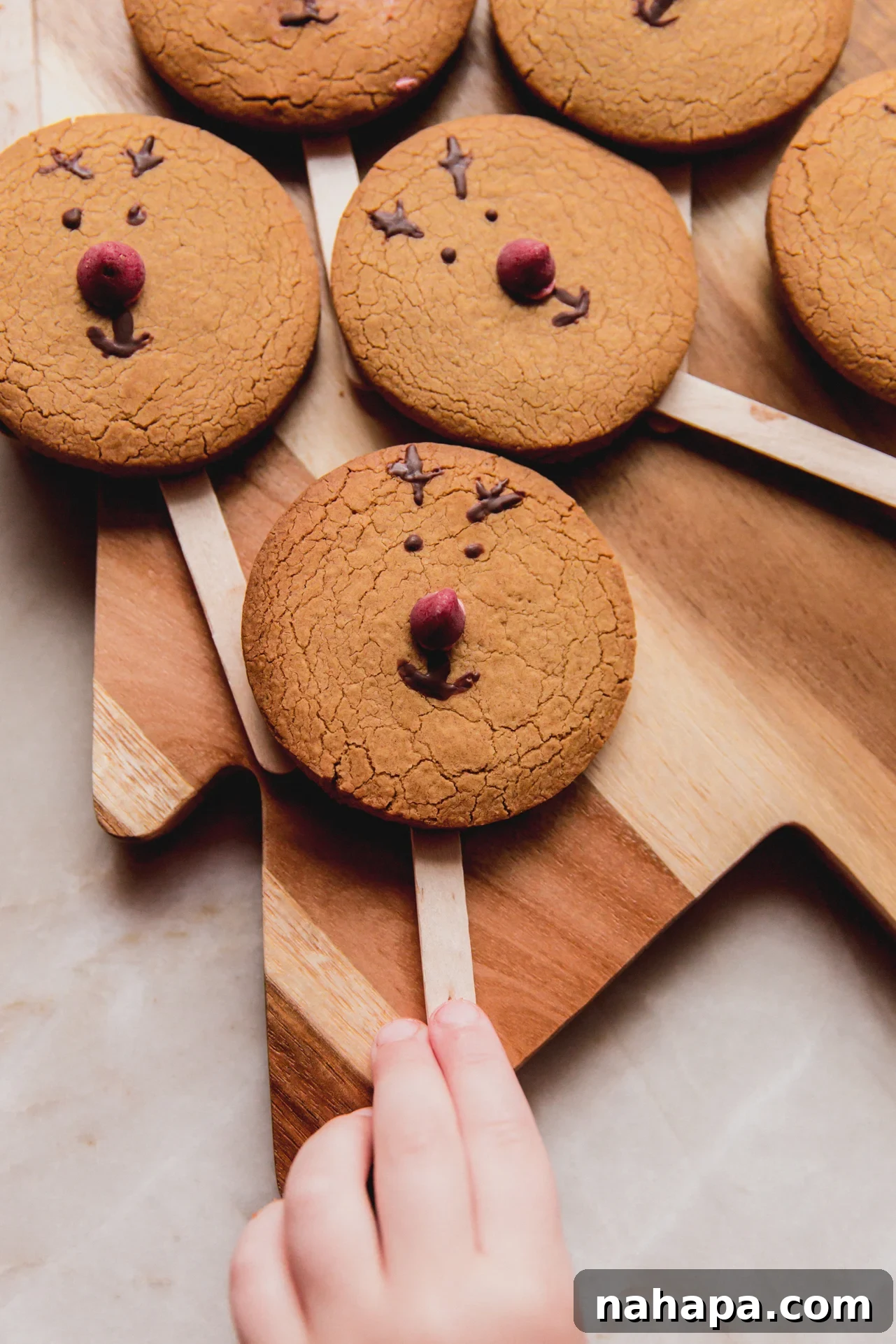 A cutting board filled with decorated gingerbread reindeer cookies, with a child's hand reaching playfully for one.