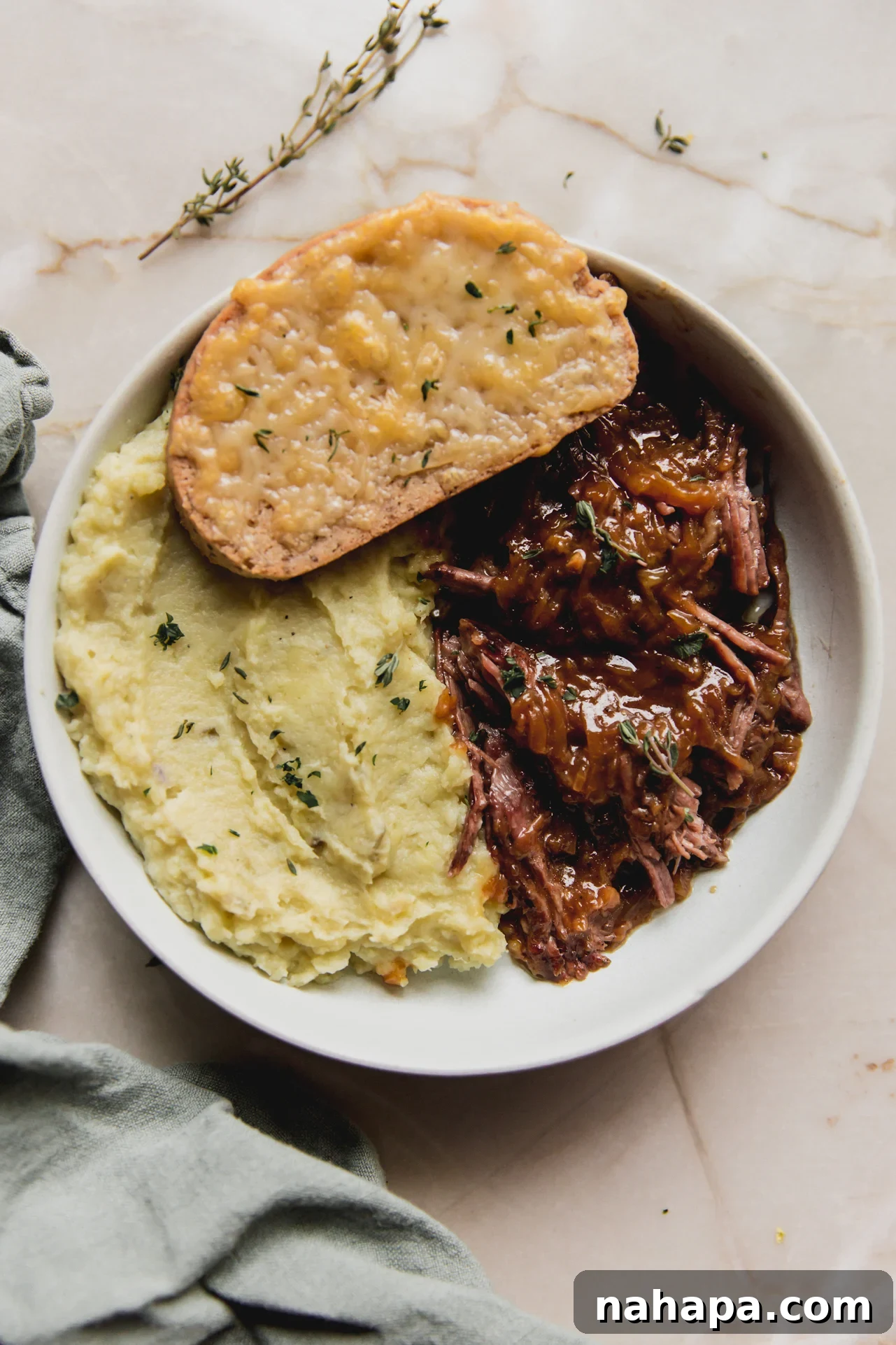 French Onion Braised Beef 11 A serving of French onion pot roast in a shallow bowl, garnished with fresh herbs, accompanied by mashed sweet potato and a piece of cheese toast.