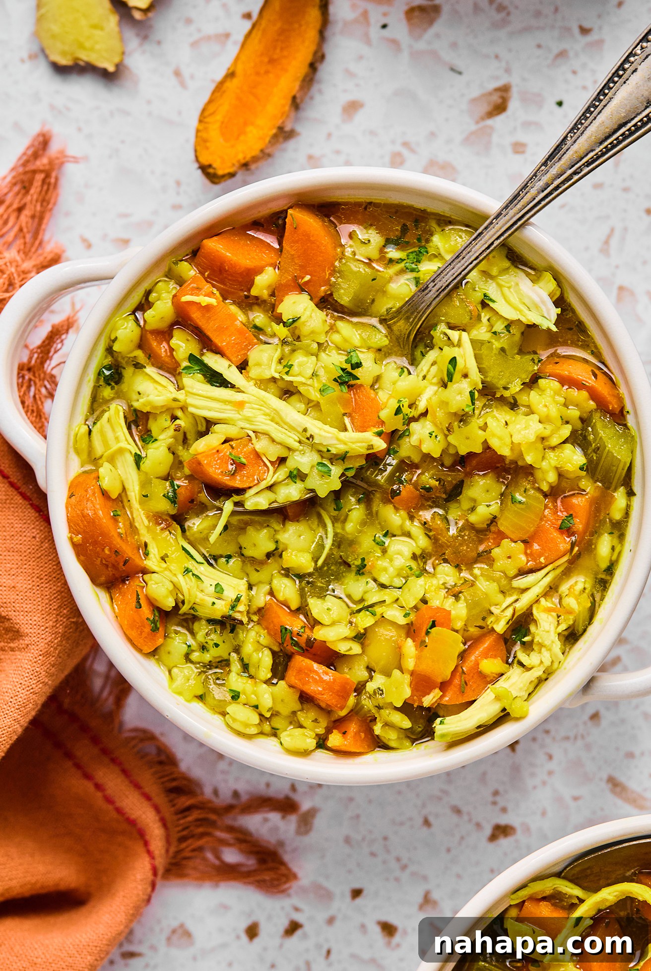 A close-up shot of a steaming bowl of Lemon Turmeric Chicken Noodle Soup, with a spoon nestled inside, inviting the viewer to enjoy its warmth and flavor.