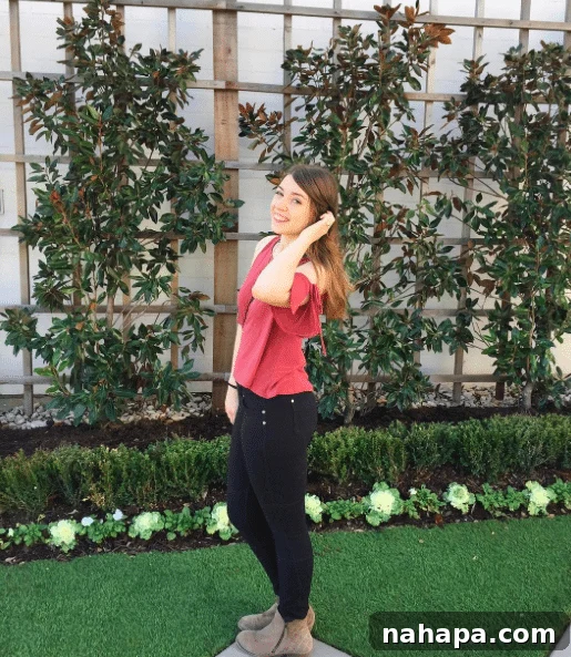A brunette woman in black pants and a pink shirt standing in front of greenery. 