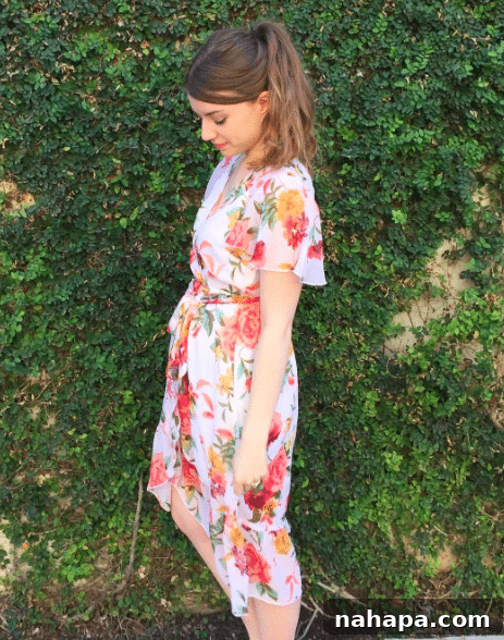 A brunette woman in a floral wrap dress standing in front of a green foliage wall.