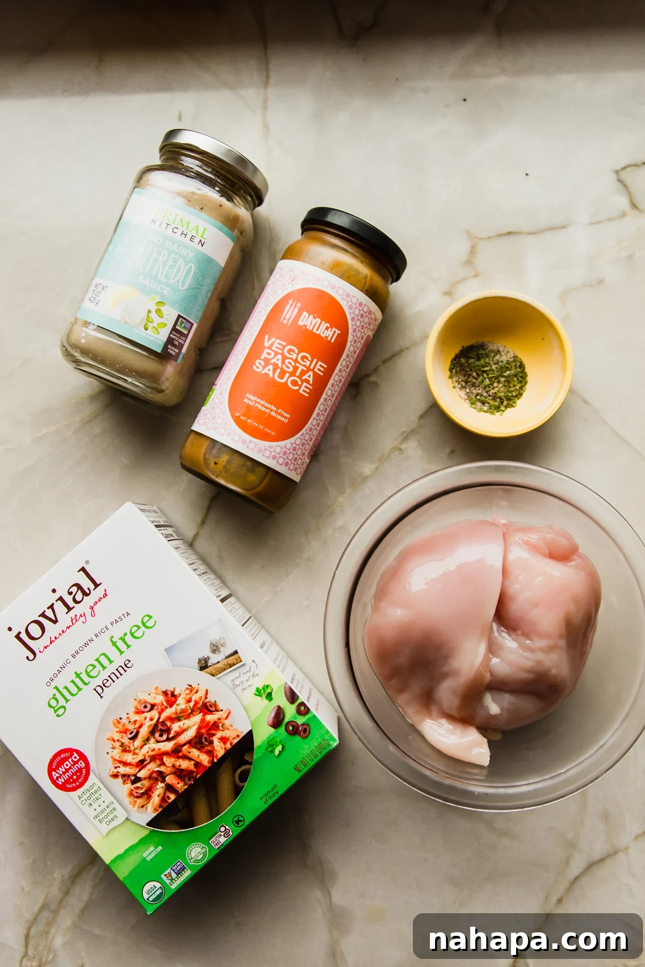 Velvety Slow Cooker Chicken Pasta 3 A selection of fresh ingredients including chicken breast, various spices, a jar of Daylight veggie pasta sauce, dairy-free alfredo sauce, and gluten-free pasta.
