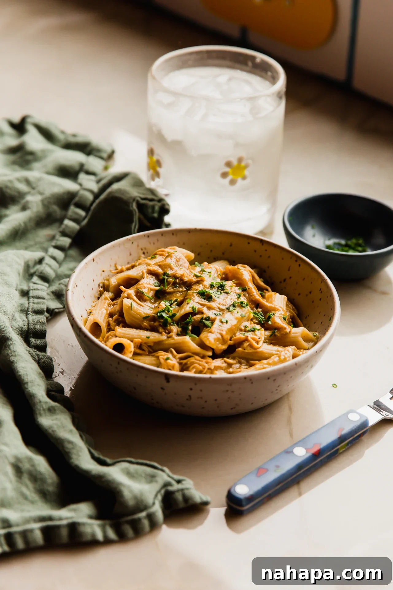 Velvety Slow Cooker Chicken Pasta 8 A close-up of creamy chicken pasta in a rustic bowl, with a glass of water and fresh herbs in the soft-focused background, emphasizing comfort and simplicity.