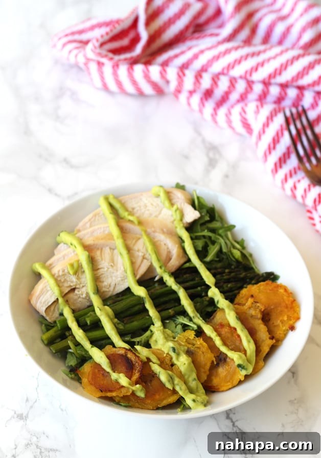 A bowl filled with fried plantains, chicken, aspargus, greens and topped with dressing.