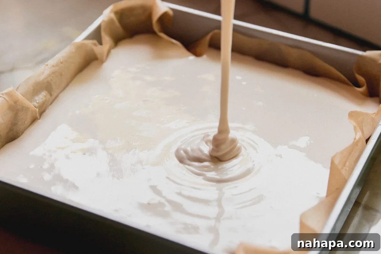Pouring the freshly whipped protein marshmallow mixture over the cooled graham cracker crust in the baking pan, showing the layering process.