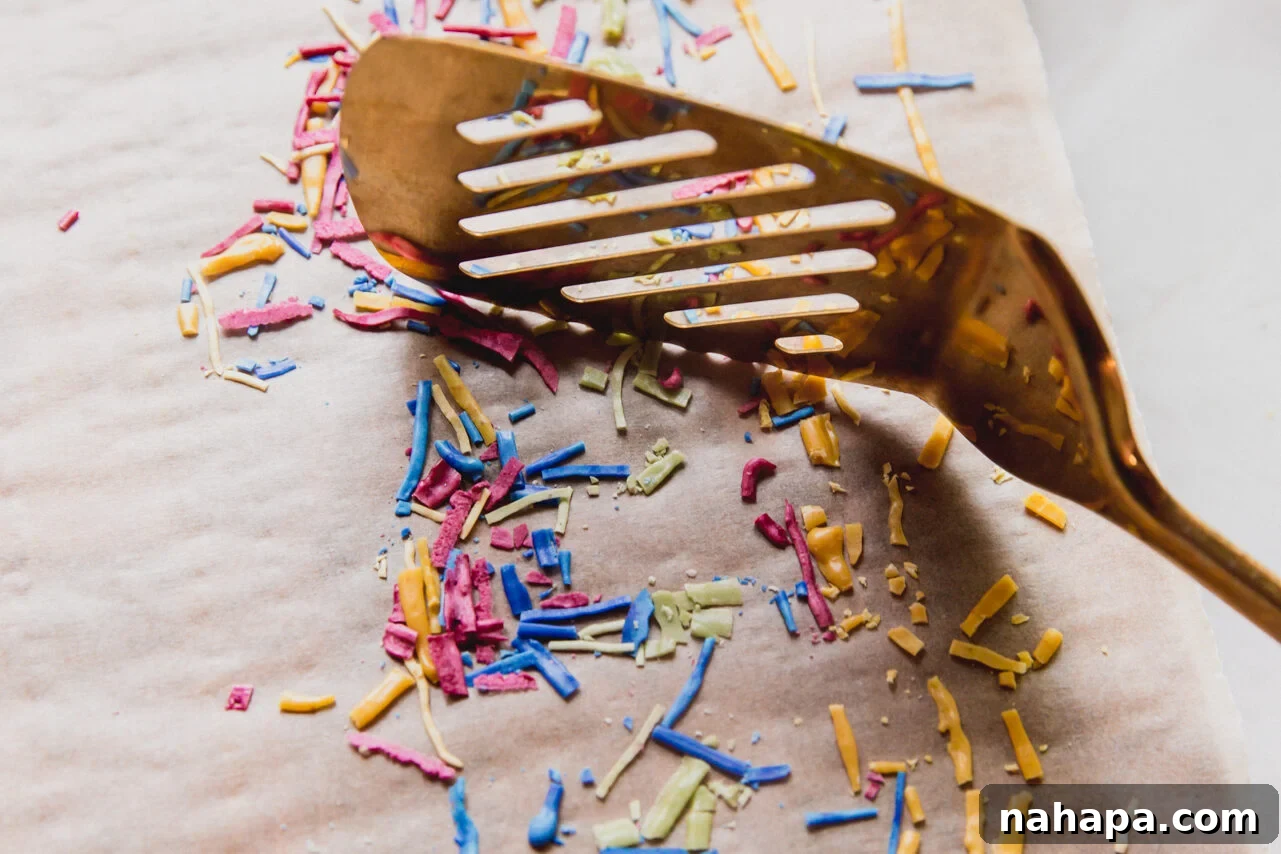 A spatula gently breaking up long, dried lines of icing into individual homemade sprinkles.