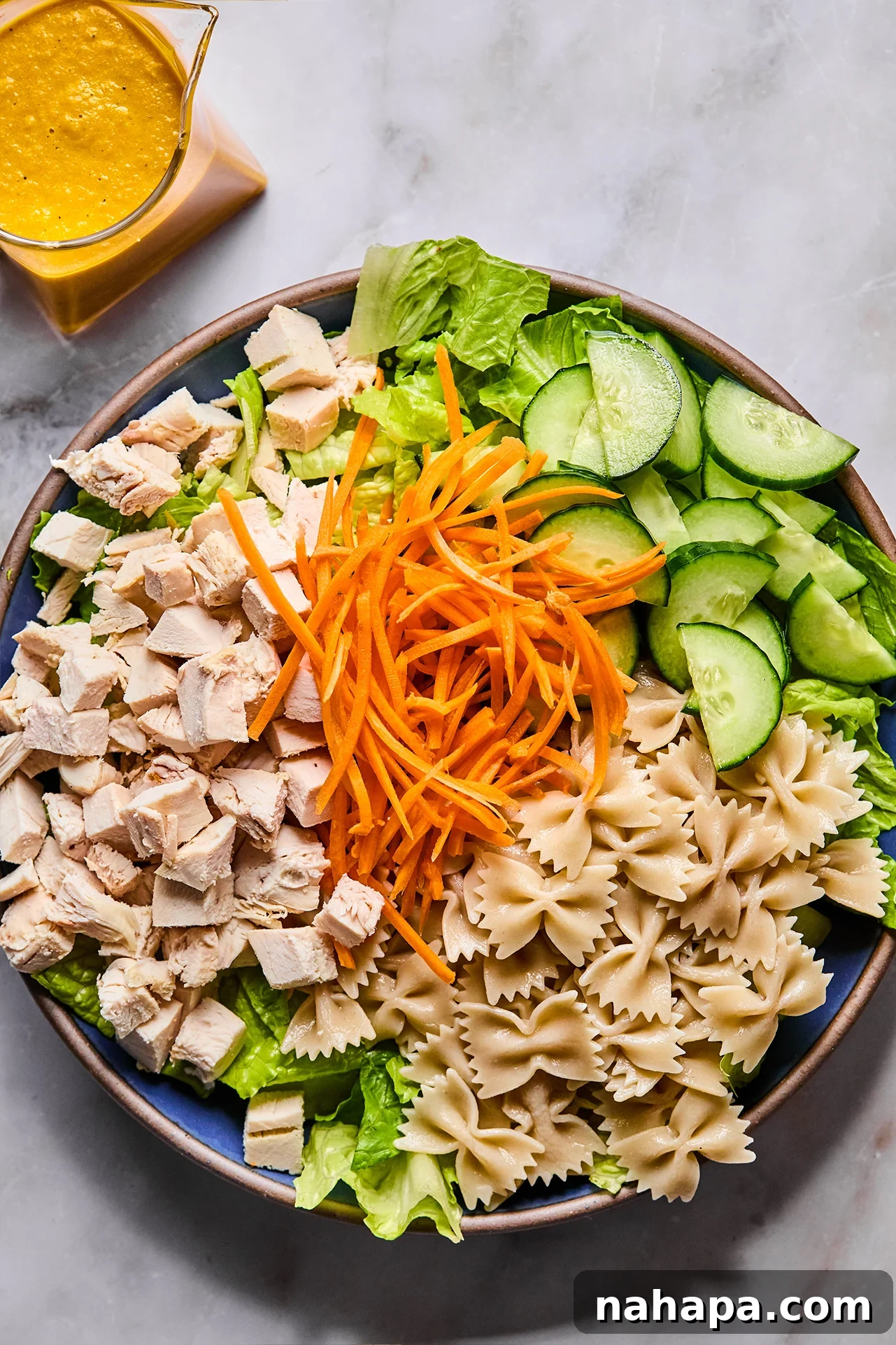 A large salad bowl filled with cooked bow-tie pasta, chopped romaine lettuce, diced cucumber, shredded carrots, and cubed chicken.