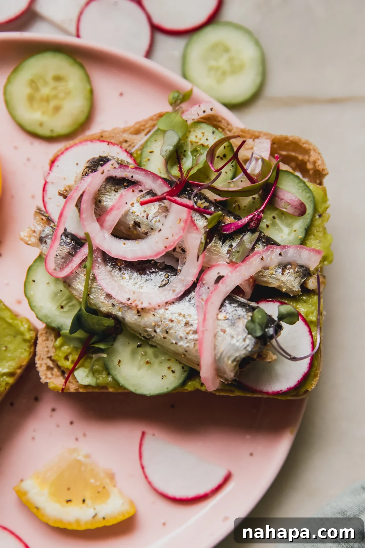 Sardine toast topped with sardines, cucumber, pickled onion and microgreens on a pink plate.