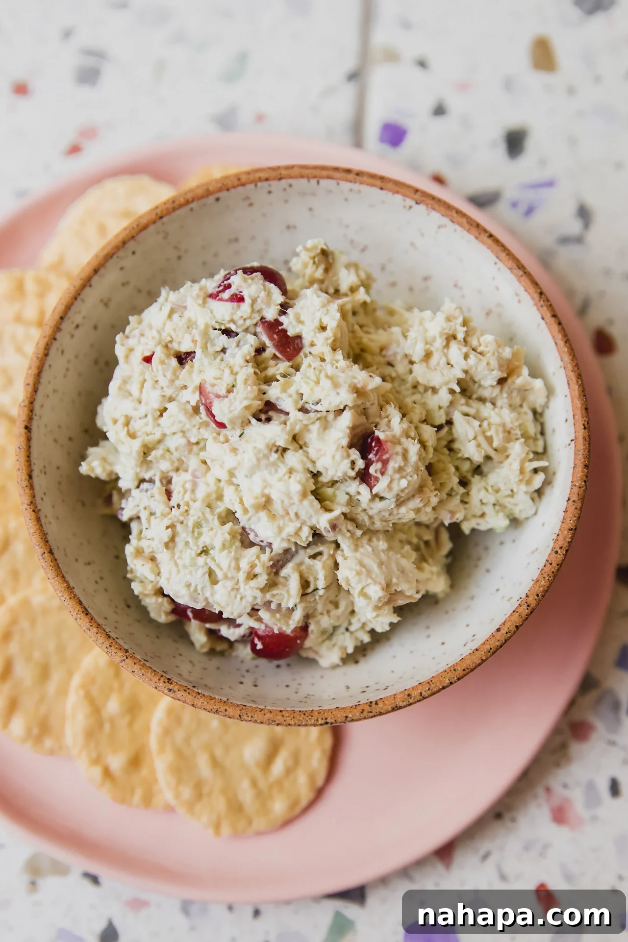 A small bowl of grape chicken salad on a pink plate, accompanied by gluten-free crackers for serving.