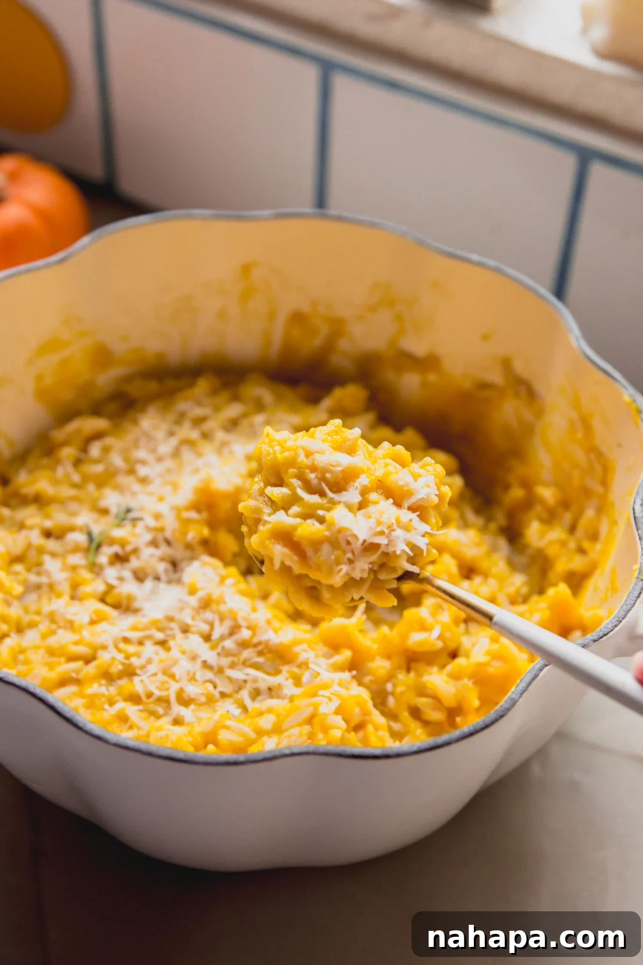 A serving spoon lifting a generous portion of creamy pumpkin orzo from a large serving dish, highlighting the inviting texture and seasonal appeal of the pasta.