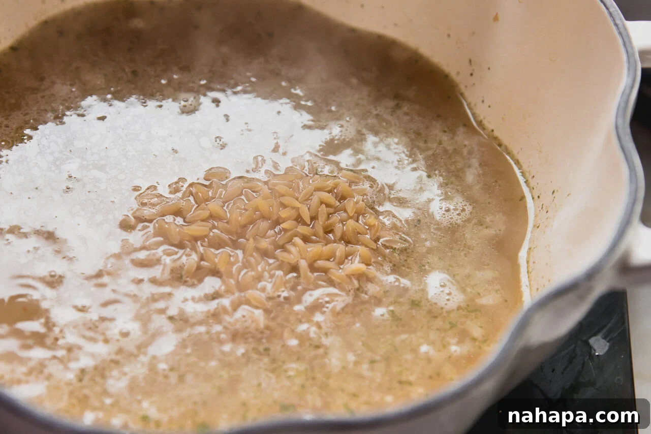 A pot on the stove containing gluten-free orzo simmering in a rich, savory liquid with aromatics.