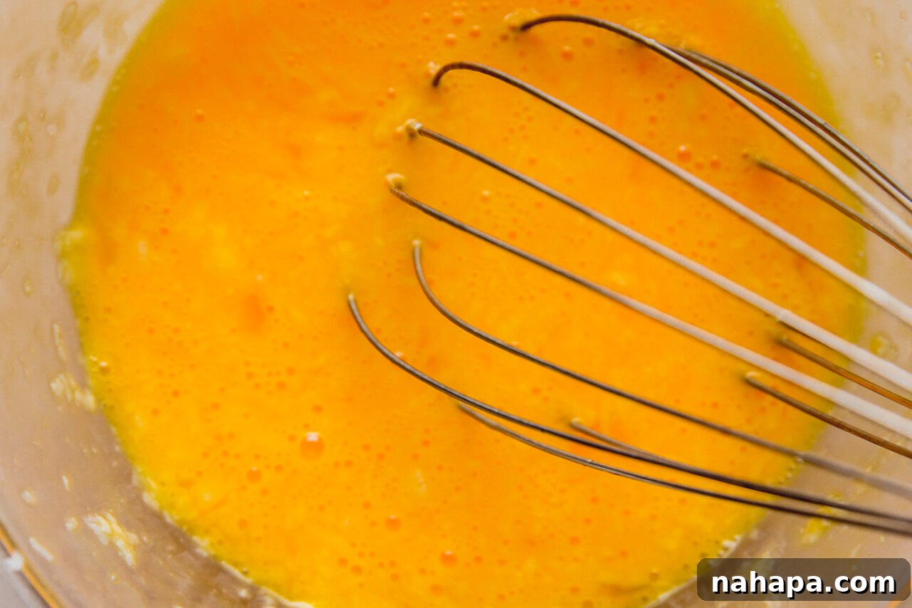 A whisk vigorously mixing eggs and grated cheese together in a glass bowl until well combined.