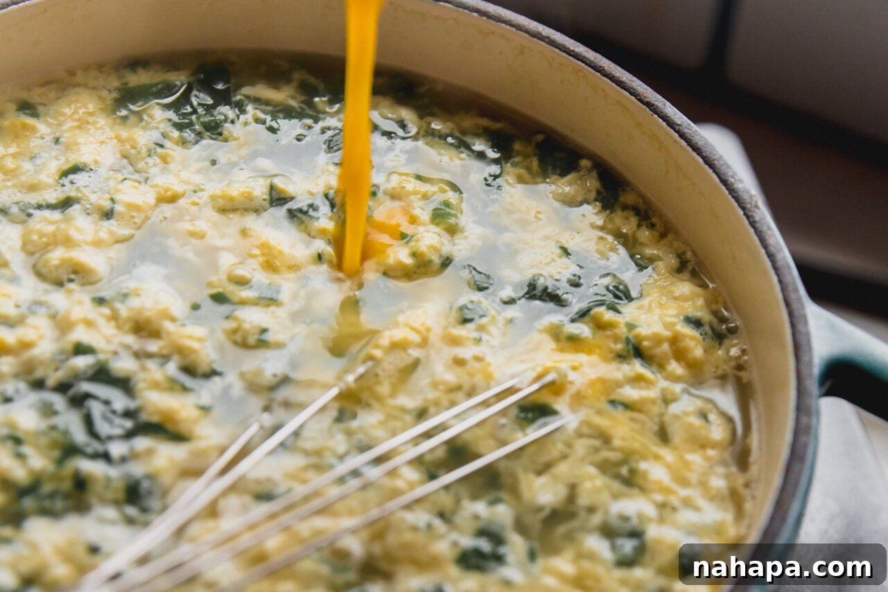 The whisked egg and cheese mixture being slowly streamed into the simmering pot of spinach and broth, creating delicate egg ribbons.