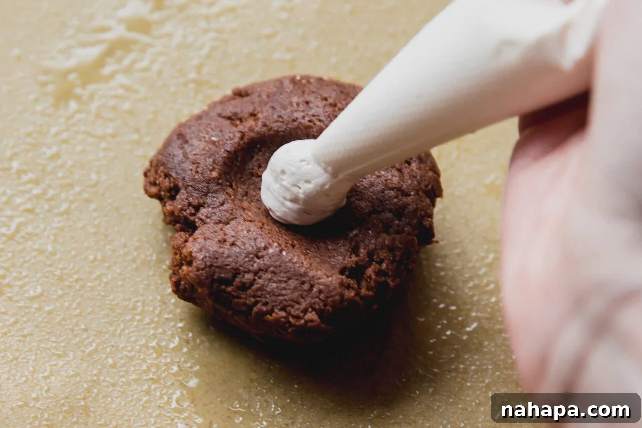 A hand expertly piping fluffy marshmallow cream into the thumbprint of a chocolate cookie dough ball on parchment paper, preparing it for baking.