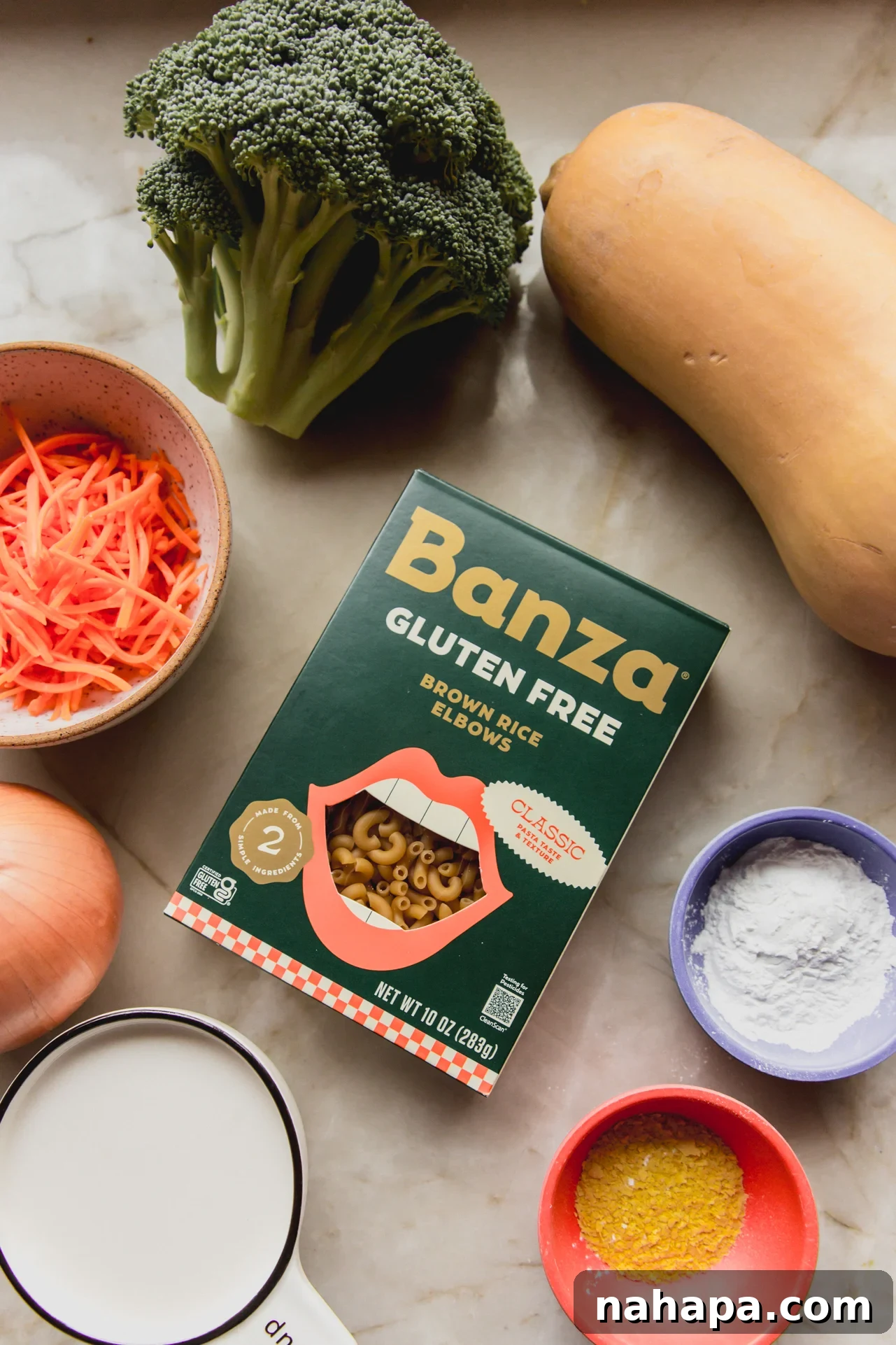 A flat lay of fresh ingredients for broccoli cheese noodle soup, including broccoli, carrots, onion, garlic, nutritional yeast, and Banza pasta, on a marble countertop.