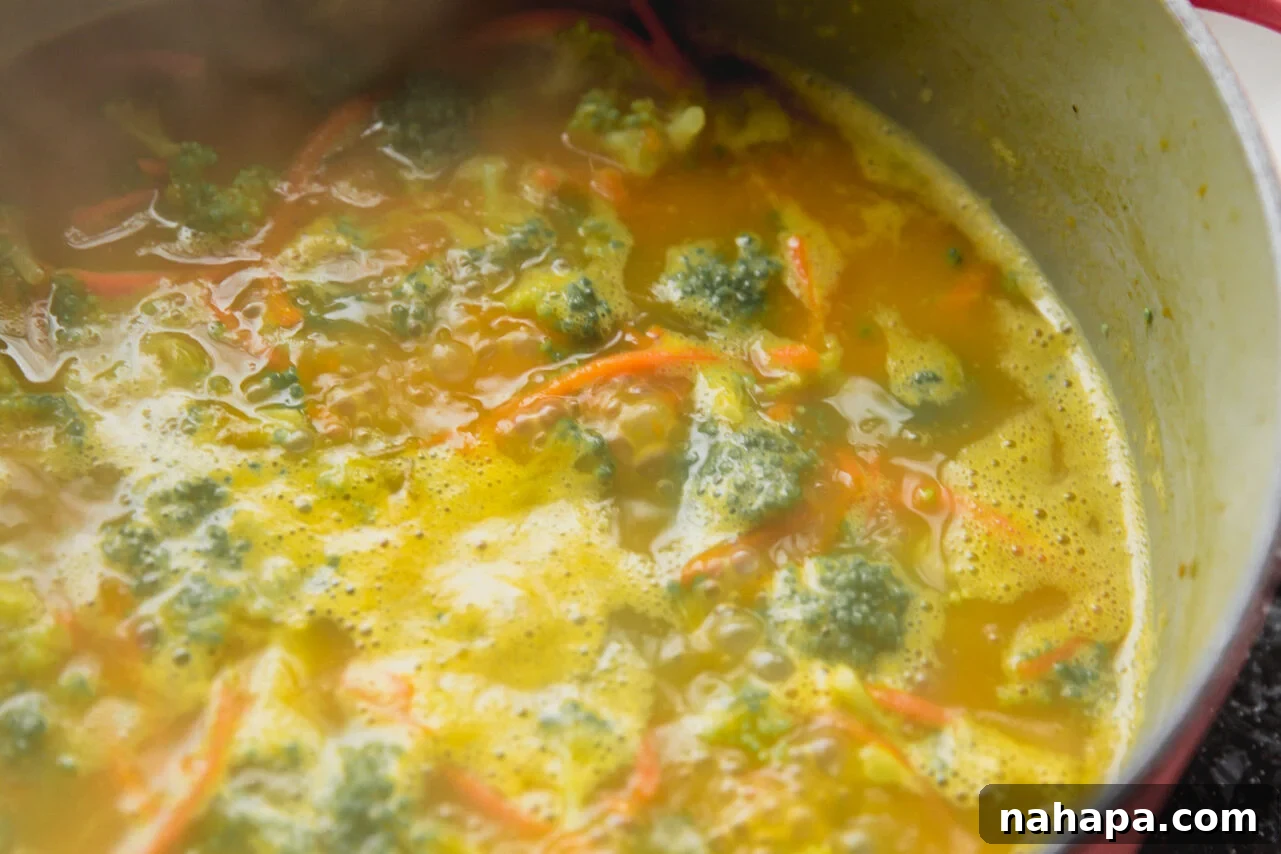 Creamy broccoli cheese soup simmering in a large pot, with broccoli florets and shredded carrots visibly integrated.