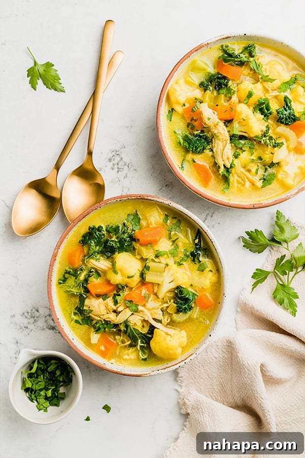 Overhead shot of two bowls of turmeric chicken soup, one garnished with cilantro and lime, on a rustic wooden table.