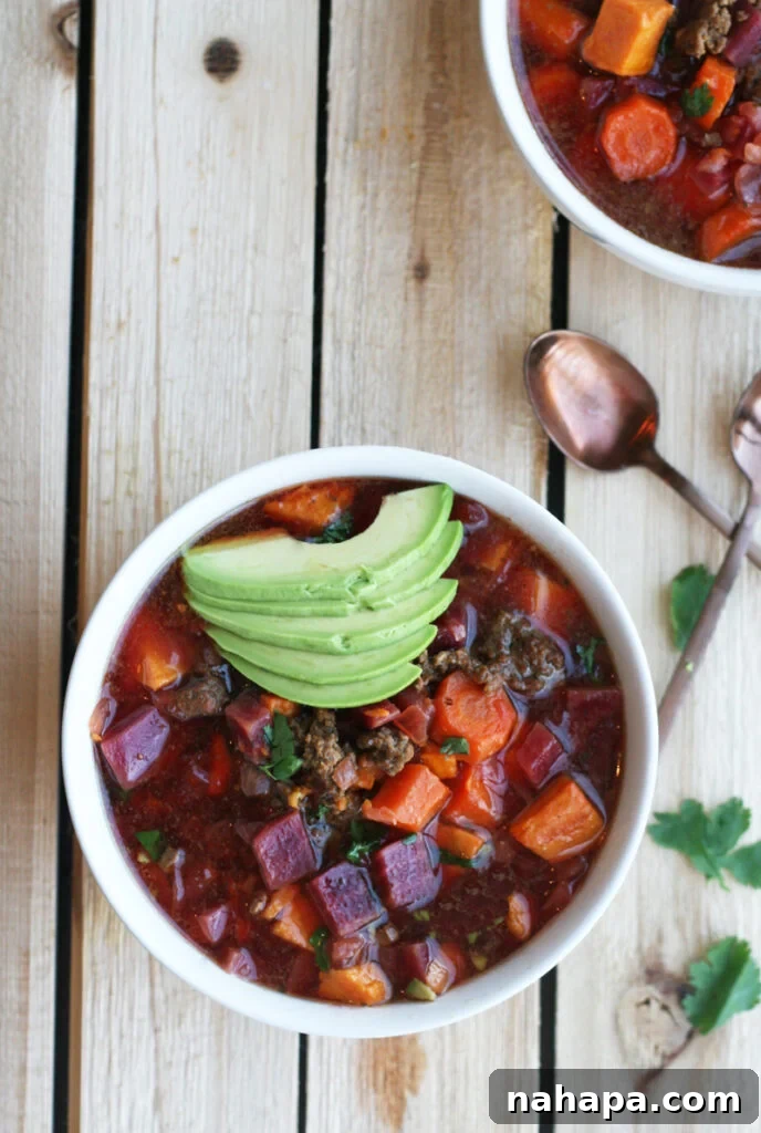 A white bowl filled with AIP chili, topped with avocado slices, on a wooden table.