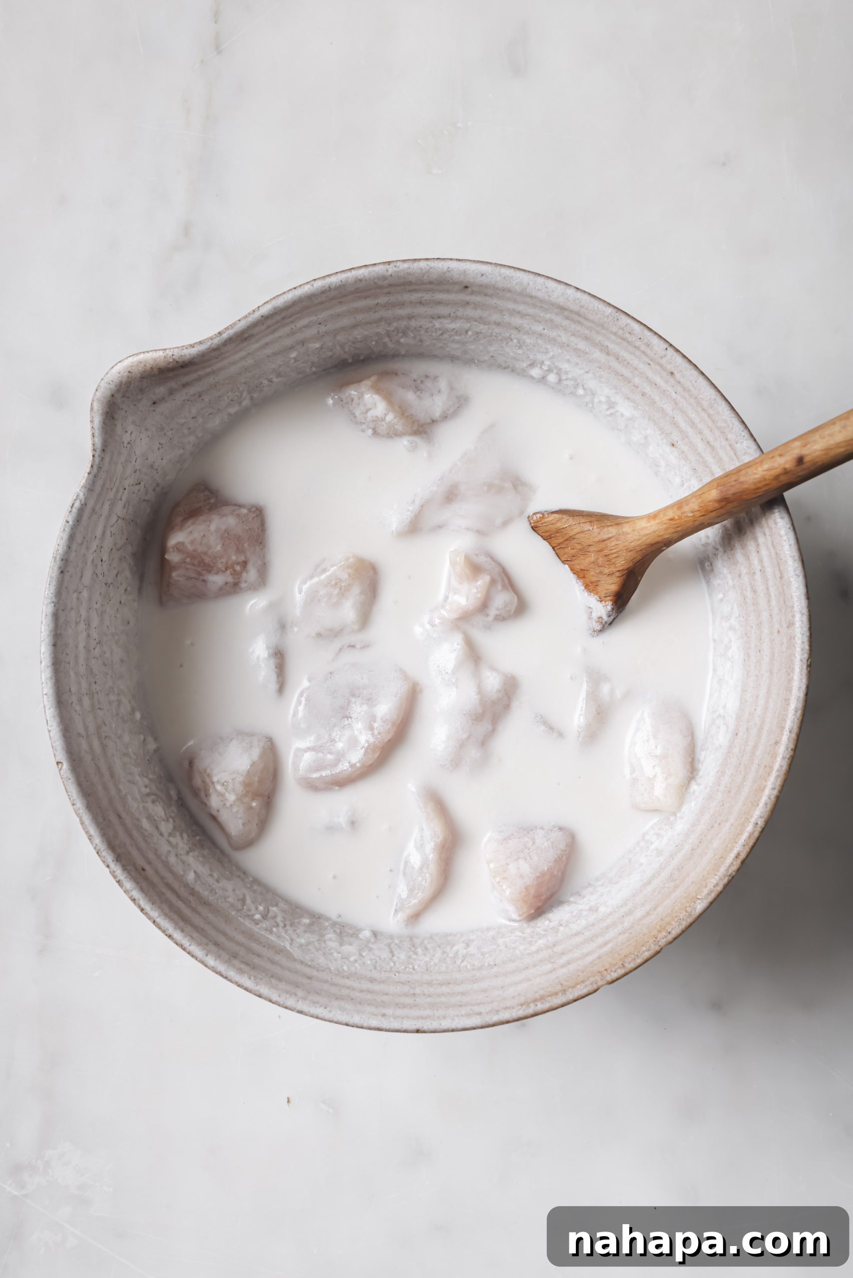 Cubed chicken breast marinating in a mixture of coconut milk and apple cider vinegar in a glass bowl.