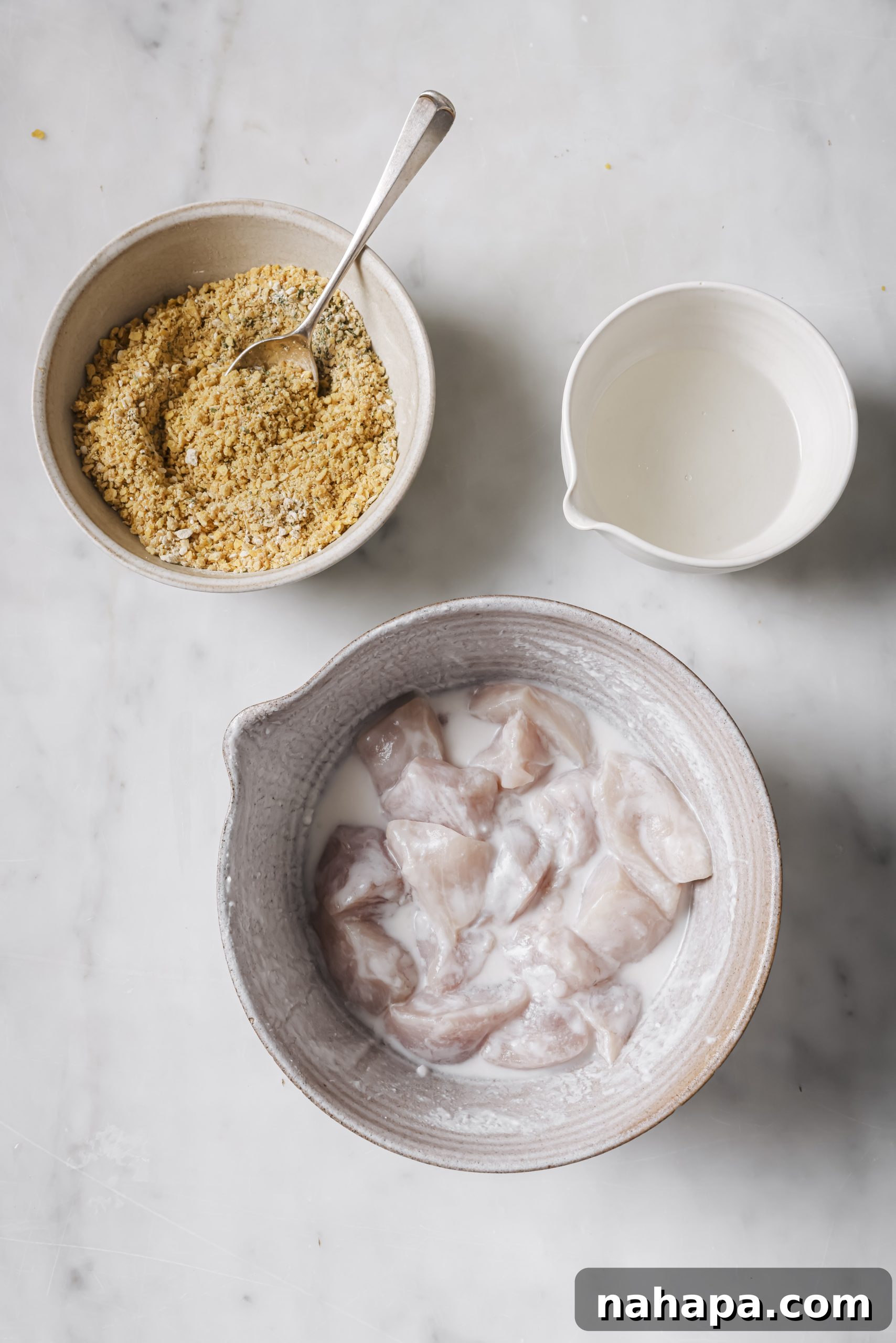 Three separate bowls: one with marinated chicken, one with ground plantain chip mixture, and one with melted coconut oil, set up for a dredging station.