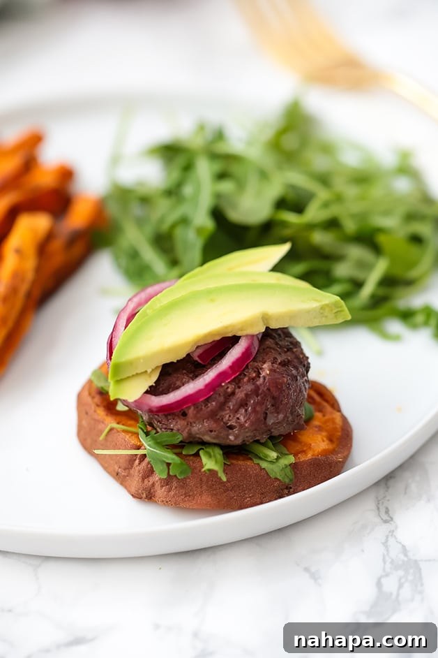 Close-up of a single beef slider on a roasted sweet potato bun, garnished with vibrant pickled onions and creamy avocado slices. Served with sweet potato fries and arugula.