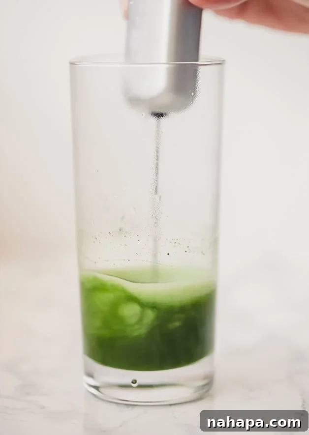 A close-up of a glass where vibrant green matcha powder and hot water are being vigorously mixed with a metal handheld milk frother, creating a frothy paste.