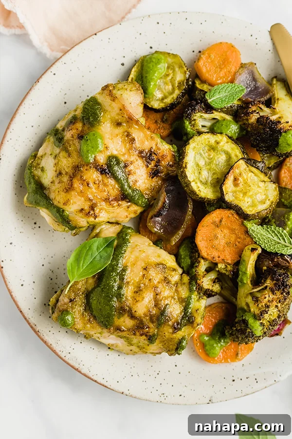 Close-up of roasted chicken and vegetables on a white plate, drizzled with fresh pesto.