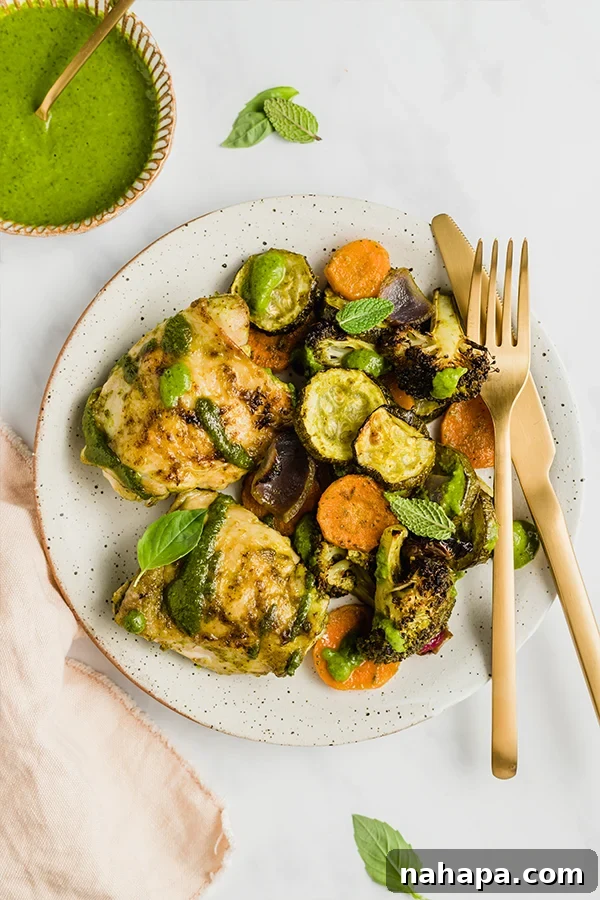 A serving of chicken pesto with roasted vegetables on a white plate, with a small bowl of green pesto on the side.