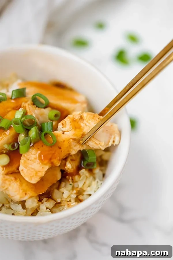 A white bowl with cauliflower rice topped with chicken teriyaki garnished with green onion, chopsticks lifting a piece of chicken.