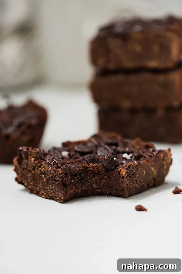 A zucchini brownie with a bite taken out of it in front of a stack of zucchini brownies.