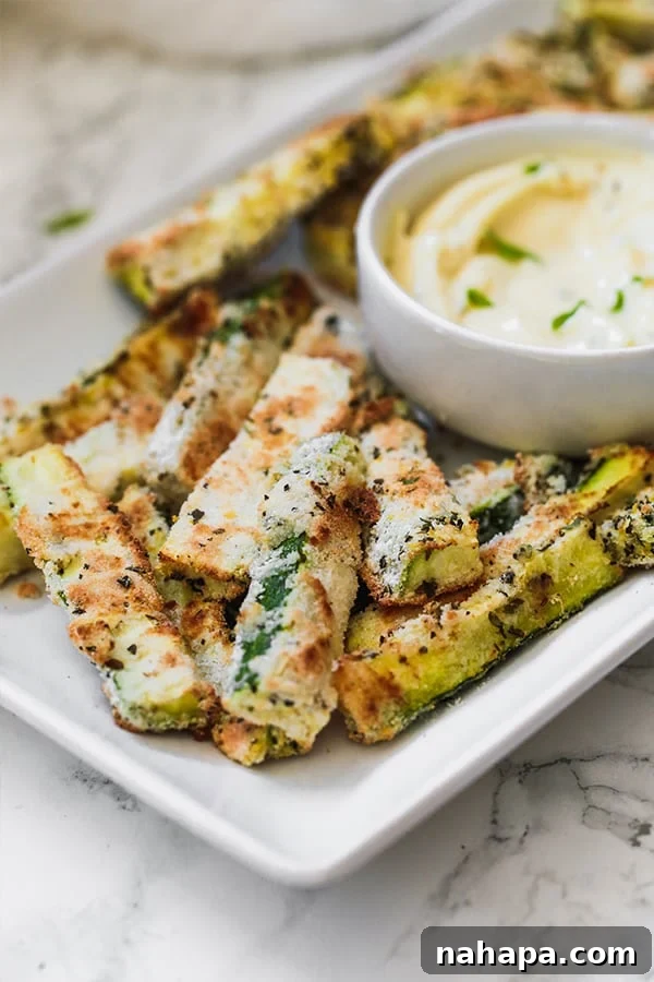 A white serving dish filled with perfectly baked zucchini fries, arranged artfully around a small white bowl of a creamy dipping sauce, ready for sharing.